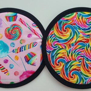 May include: Two round fabric coasters with black trim. One coaster features a pink background with candy illustrations, including lollipops, cotton candy, and cupcakes. The other coaster has a colorful swirl design.