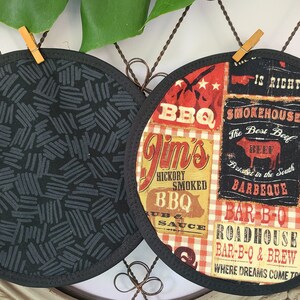 May include: Two round, black and gray fabric pot holders. One features a black scribble pattern, the other a collage of BBQ-themed text and graphics in red, white, and black. The pot holders are trimmed with black binding.