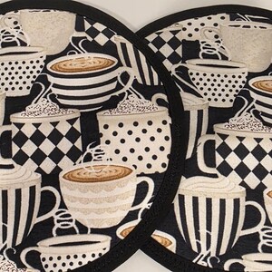 May include: Two round black and white patterned pot holders. The design features various coffee cups with striped, polka dot, and checkered patterns. The cups are filled with coffee and topped with whipped cream. The pot holders have a black border.