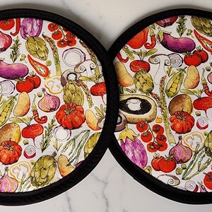 May include: Two round, fabric pot holders with a colorful vegetable pattern. The design includes tomatoes, eggplants, artichokes, and mushrooms. Each pot holder has a black border.