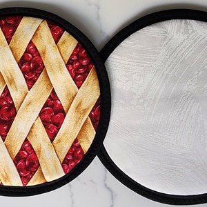 Peut inclure: Deux dessous de plat ronds en tissu. L'un présente un motif de tarte aux cerises avec une croûte en treillis et une garniture de cerises rouges. L'autre a un motif abstrait blanc et gris. Les deux ont une bordure noire.