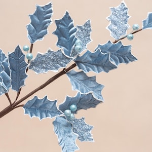 May include: A decorative artificial holly branch featuring light blue leaves with white edges and silver glitter accents. The branch includes small, round, light blue berries attached to a brown stem, creating a textured, wintry appearance.