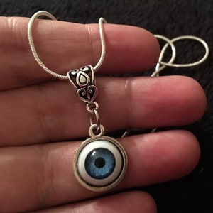 Puede incluir: Un collar de cadena de plata con un colgante de plata que presenta un ojo azul con una pupilla negra. El ojo está colocado en un círculo blanco con un borde plateado.