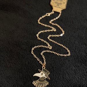K&ouml;nnte beinhalten: Eine goldene Kettenkette mit einem schwarzen und wei&szlig;en Emaille-Anh&auml;nger, der einen Pestarzt mit einer langen Schnabelmaske und einem Hut darstellt.