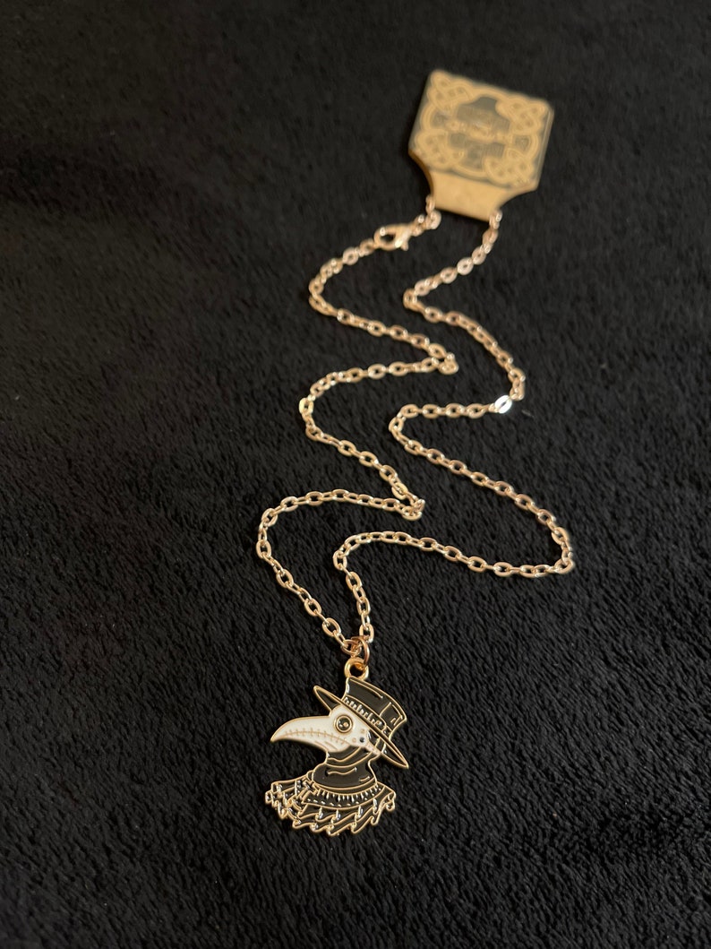 K&ouml;nnte beinhalten: Eine goldene Kettenkette mit einem schwarzen und wei&szlig;en Emaille-Anh&auml;nger, der einen Pestarzt mit einer langen Schnabelmaske und einem Hut darstellt.