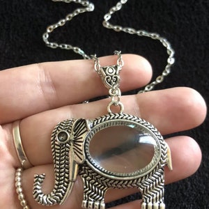 Puede incluir: Un collar con un colgante de elefante de plata con una lupa en el centro. El elefante está detallado con un patrón texturizado y tiene una lupa de vidrio transparente.