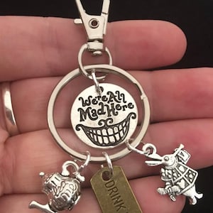 Puede incluir: Llavero de plata con un dije redondo que dice "We're All Mad Here" con una sonrisa del Gato de Cheshire. El llavero también tiene tres dijes: una tetera de plata, un conejo de plata y una etiqueta de bronce que dice "Drink Me."