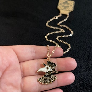 K&ouml;nnte beinhalten: Eine goldene Kettenkette mit einem schwarzen und wei&szlig;en Emaille-Anh&auml;nger, der einen Pestarzt mit einer langen Schnabelmaske und einem Hut darstellt.