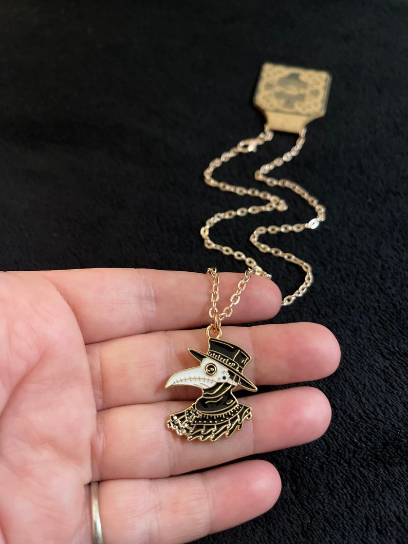 K&ouml;nnte beinhalten: Eine goldene Kettenkette mit einem schwarzen und wei&szlig;en Emaille-Anh&auml;nger, der einen Pestarzt mit einer langen Schnabelmaske und einem Hut darstellt.