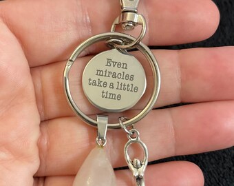 May include: Silver keychain with a charm that reads "Even miracles take a little time". The keychain also has a pink crystal charm and a silver charm of a woman with a spiral.