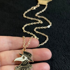 K&ouml;nnte beinhalten: Eine goldene Kettenkette mit einem schwarzen und wei&szlig;en Emaille-Anh&auml;nger, der einen Pestarzt darstellt, der einen langen schwarzen Mantel, einen Hut und eine Schnabelmaske tr&auml;gt.