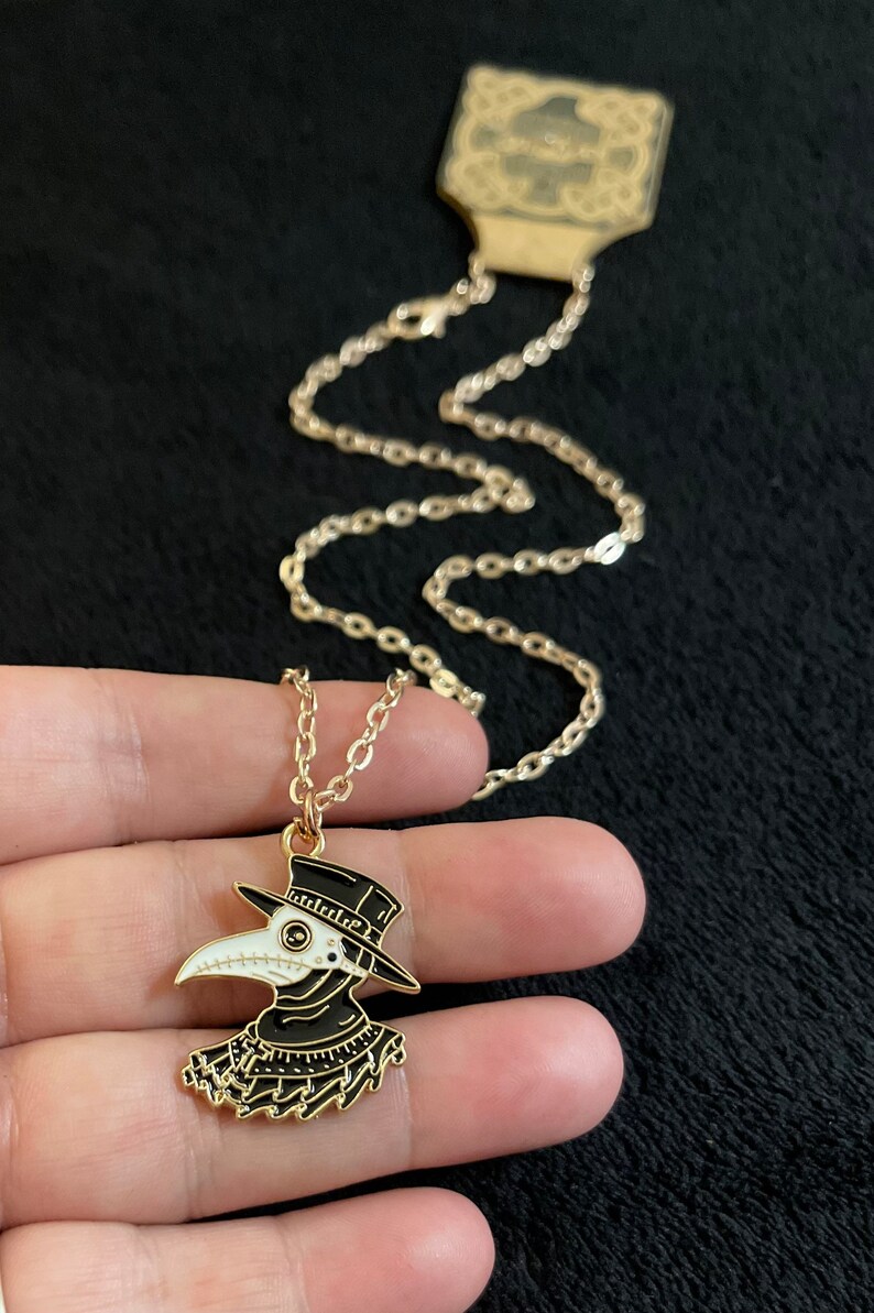 K&ouml;nnte beinhalten: Eine goldene Kettenkette mit einem schwarzen und wei&szlig;en Emaille-Anh&auml;nger, der einen Pestarzt darstellt, der einen langen schwarzen Mantel, einen Hut und eine Schnabelmaske tr&auml;gt.