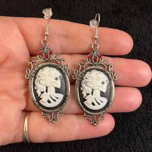 Puede incluir: Un par de pendientes colgantes plateados. Cada pendiente presenta un camafeo ovalado con un diseño de esqueleto blanco sobre un fondo negro. Los pendientes tienen detalles rojos y filigranas decorativas.