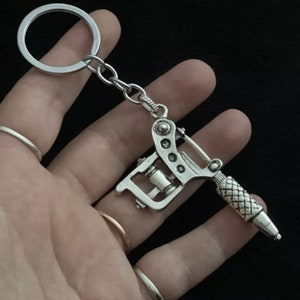 Puede incluir: Llavero plateado con una máquina de tatuaje en miniatura. El llavero tiene un diseño detallado, incluyendo la aguja y el agarre, y está unido a una cadena y un llavero redondo. El fondo es de color negro sólido.