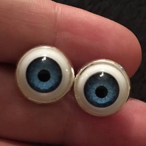 Op de afbeelding: Een paar zilveren manchetknopen met blauwe oogontwerpen. De manchetknopen zijn rond en hebben een witte achtergrond met een blauwe iris en een zwarte pupil.