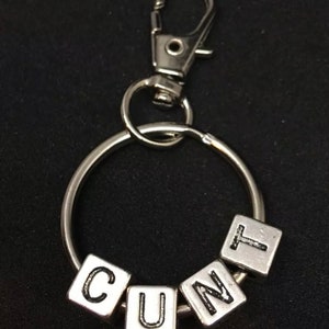 May include: Silver keyring with a silver ring and a silver clip. The ring has four silver letter blocks that spell out the word "CUNT".