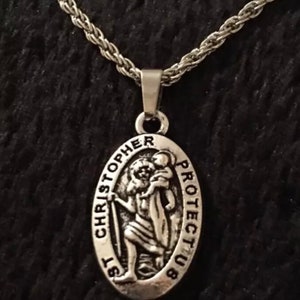 Könnte beinhalten: Silberne Kettenkette mit einem silbernen ovalen Anhänger, der eine Darstellung des Heiligen Christophorus zeigt, der ein Kind trägt. Der Anhänger trägt die Inschrift "St Christopher Protect Us".