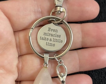 May include: Silver keychain with a round charm that reads "Even miracles take a little time". The keychain also has a pink teardrop-shaped charm and a silver charm of a woman with a spiral design.