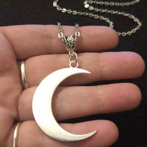 Puede incluir: Un collar con un colgante de media luna de plata en una cadena de plata. El colgante es grande y cuelga de una cadena delicada.