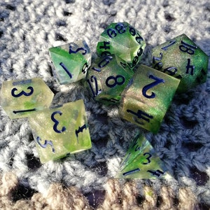 May include: A set of ten-sided dice with green and white speckled paint and blue numbers. The dice are on a gray crocheted surface.
