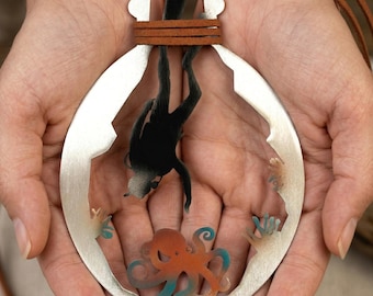 May include: A silver pendant necklace depicting a diver and an octopus inside a circular frame. The diver is black, and the octopus is orange with blue tentacles. The necklace has a brown leather cord.