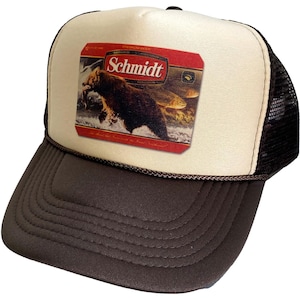 May include: A brown and beige trucker hat with a brown mesh back. The front of the hat features a vintage-style label with a brown bear catching a fish. The label reads "Schmidt" in red letters.