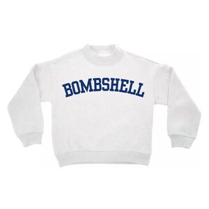 May include: A white long-sleeve sweatshirt with a mock neck collar and ribbed cuffs. The word BOMBSHELL is printed across the chest in a dark blue, arched, collegiate-style font. The garment features a simple, clean design with long sleeves.