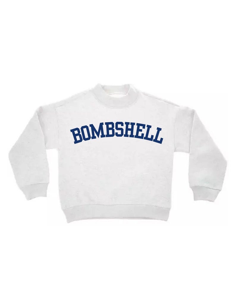 May include: A white long-sleeve sweatshirt with a mock neck collar and ribbed cuffs. The word BOMBSHELL is printed across the chest in a dark blue, arched, collegiate-style font. The garment features a simple, clean design with long sleeves.