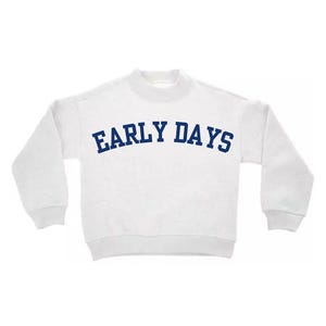 May include: A white long-sleeve sweatshirt featuring a mock neck collar and ribbed cuffs. The text "EARLY DAYS" is printed across the chest in a bold, arched, dark blue font. The garment has a simple, clean design with long sleeves and a straight hem.