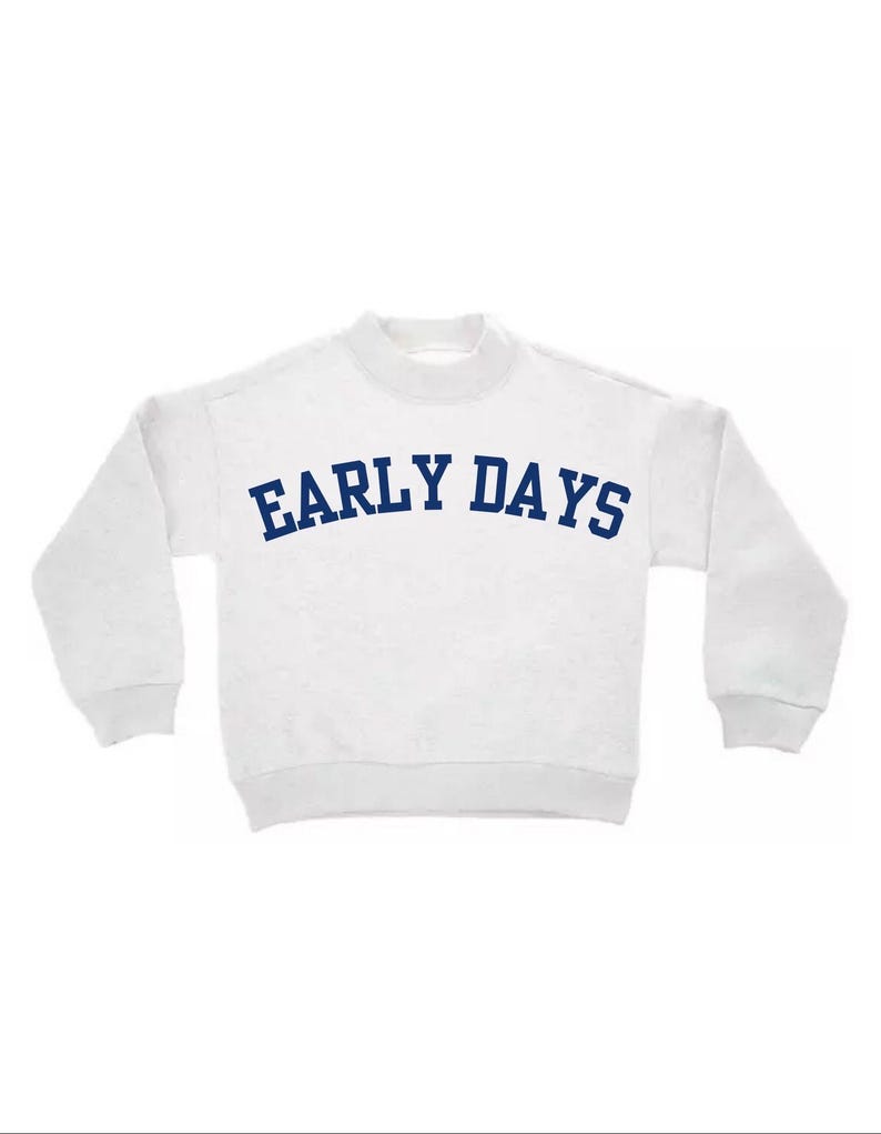May include: A white long-sleeve sweatshirt featuring a mock neck collar and ribbed cuffs. The text "EARLY DAYS" is printed across the chest in a bold, arched, dark blue font. The garment has a simple, clean design with long sleeves and a straight hem.