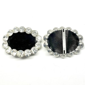 May include: Two silver-toned metal belt buckles with a black leather backing. One buckle is adorned with a circle of clear rhinestones.