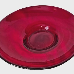 May include: A circular glass plate in a deep, translucent ruby red color. The plate features a smooth, polished surface with a slightly concave center and a wide, gently sloping rim, showcasing a consistent, vibrant red hue throughout the glass.