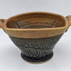 May include: A handcrafted ceramic bowl with a unique design. The bowl features a light brown rim and handles, with a textured, dark blue and brown lower section. The bowl is approximately 6 inches wide and 4 inches tall, perfect for serving.