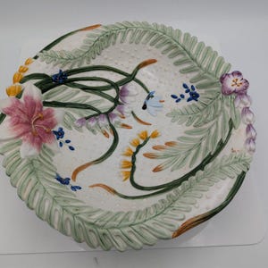 May include: A decorative ceramic plate with a white background, featuring a floral design. The plate has a scalloped edge with green fern-like leaves and colorful flowers in pink, purple, yellow, and blue. The plate has a textured surface.