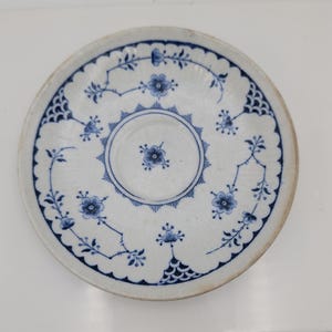 May include: A round ceramic saucer featuring a white base with blue floral patterns and intricate scalloped edges. The center has a circular indentation surrounded by a sunburst design, with delicate blue vines and net motifs decorating the rim.