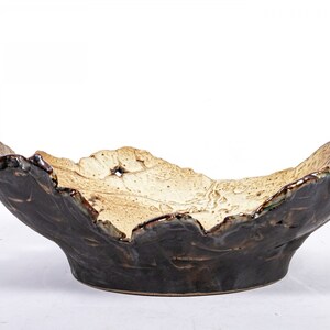 May include: A ceramic bowl with an irregular, organic shape. The bowl is glazed in a dark brown color with a lighter brown and white design on the inside. The bowl is perfect for serving snacks or fruit.