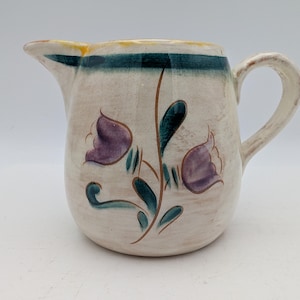 May include: Cream-colored ceramic pitcher with a curved handle and spout. The pitcher features a floral design with purple flowers and green leaves. A teal and yellow band decorates the top rim.