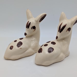 May include: A pair of off-white ceramic deer figurines in a reclining position. Each deer features hand-painted dark brown spots on its back and sides, along with dark brown accents on the ears, eyes, and forehead, creating a spotted pattern.