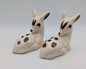 May include: A pair of off-white ceramic deer figurines in a reclining position. Each deer features hand-painted dark brown spots on its back and sides, along with dark brown accents on the ears, eyes, and forehead, creating a spotted pattern.