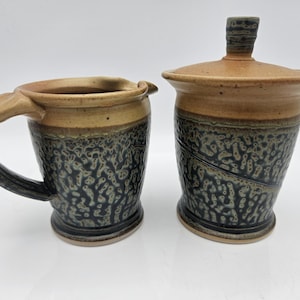 May include: A ceramic mug with a handle and a lidded jar. Both pieces have a two-tone design, with a tan upper section and a dark blue and green textured lower section. The mug has a unique handle design.