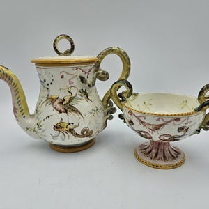 May include: A white ceramic teapot and sugar bowl with a whimsical design. The teapot has a snake-like spout and handle, while the sugar bowl has two snake-like handles. Both pieces are decorated with floral and bird motifs in shades of pink, yellow, and green.