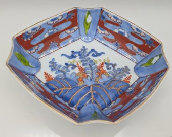 May include: A hexagonal ceramic bowl featuring a white interior with a central blue and orange botanical illustration. The rim is decorated with alternating panels of blue and red patterns, accented with small green leaf motifs and gold trim.
