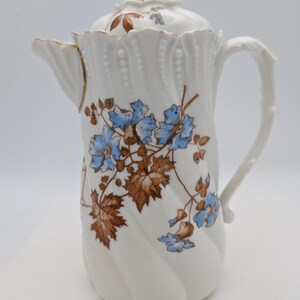 May include: White porcelain coffee pot with a lid, featuring a floral design in blue and brown. The pot has a twisted body, a curved handle, and a spout. The floral pattern includes blue flowers and brown leaves.