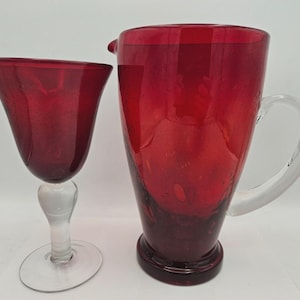 May include: A deep red glass pitcher and a matching stemmed glass goblet. Both items feature a vibrant crimson hue with visible air bubbles trapped within the glass. The pitcher has a clear glass handle, and the goblet has a clear stem and base.