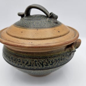 May include: A handmade ceramic lidded serving dish. The dish features a tan and dark green color scheme with a textured surface. The lid has a handle and the dish has two side handles. This piece is ideal for serving food.