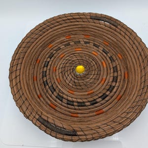 May include: A round, woven basket with a spiral design. The basket is made of brown material with black and orange accents. A yellow bead is in the center of the basket.