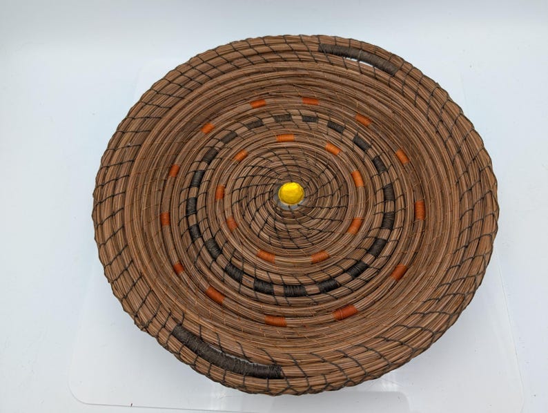 May include: A round, woven basket with a spiral design. The basket is made of brown material with black and orange accents. A yellow bead is in the center of the basket.