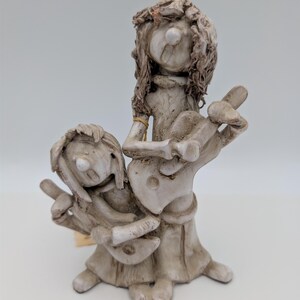May include: A light gray, textured ceramic figurine depicting two figures standing together. The taller figure has long, wavy hair and plays a guitar, while the shorter figure stands beside them, also playing a guitar. Both figures have open mouths.