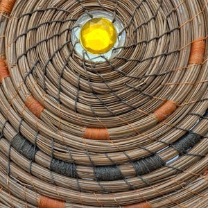 May include: Close-up of a handcrafted woven basket with a spiral design. The basket features natural brown fibers, accented with orange and dark gray stitching. A yellow gem is set in the center.