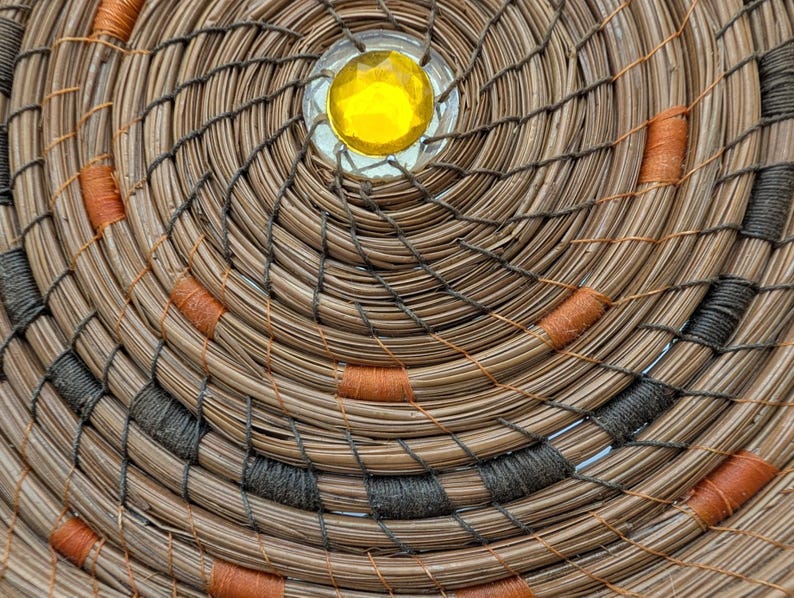 May include: Close-up of a handcrafted woven basket with a spiral design. The basket features natural brown fibers, accented with orange and dark gray stitching. A yellow gem is set in the center.